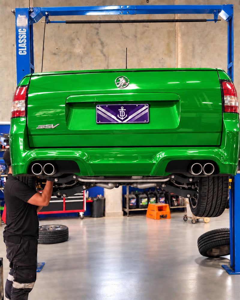 Rear of a green Holden VE SS ute on the hoist with a mechanic underneath