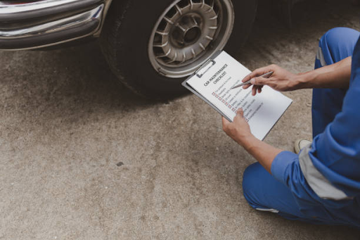 A man in blue overalls sits beside a car with a clipboard, focused on identifying necessary brake repairs for the vehicle.